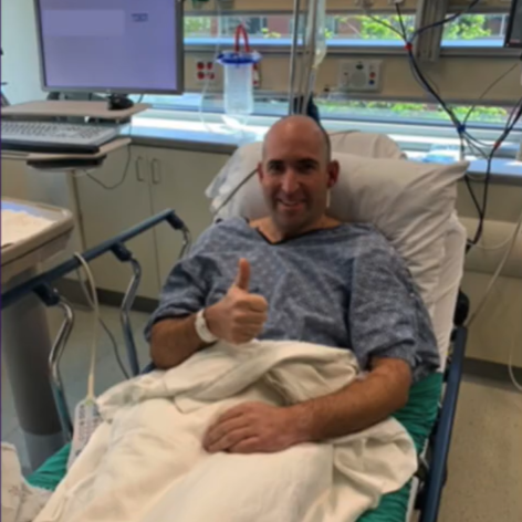 A man in a hospital bed wearing a hospital gown and covered with a white blanket gives a thumbs-up to the camera, smiling, in a medical room with monitors and equipment in the background.