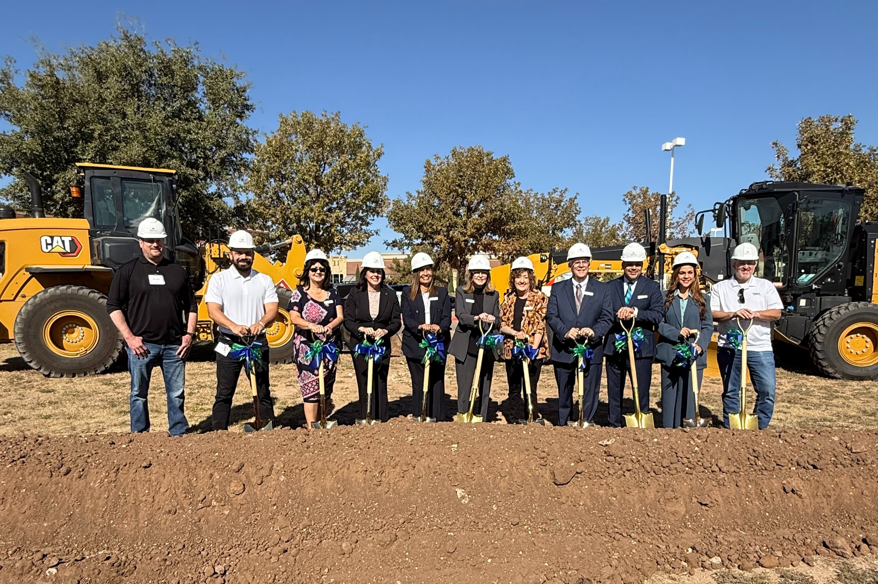 Group of people wearing hard hats and holding shovels with ribbon bows, standing in front of large construction equipment during a groundbreaking ceremony on a sunny day